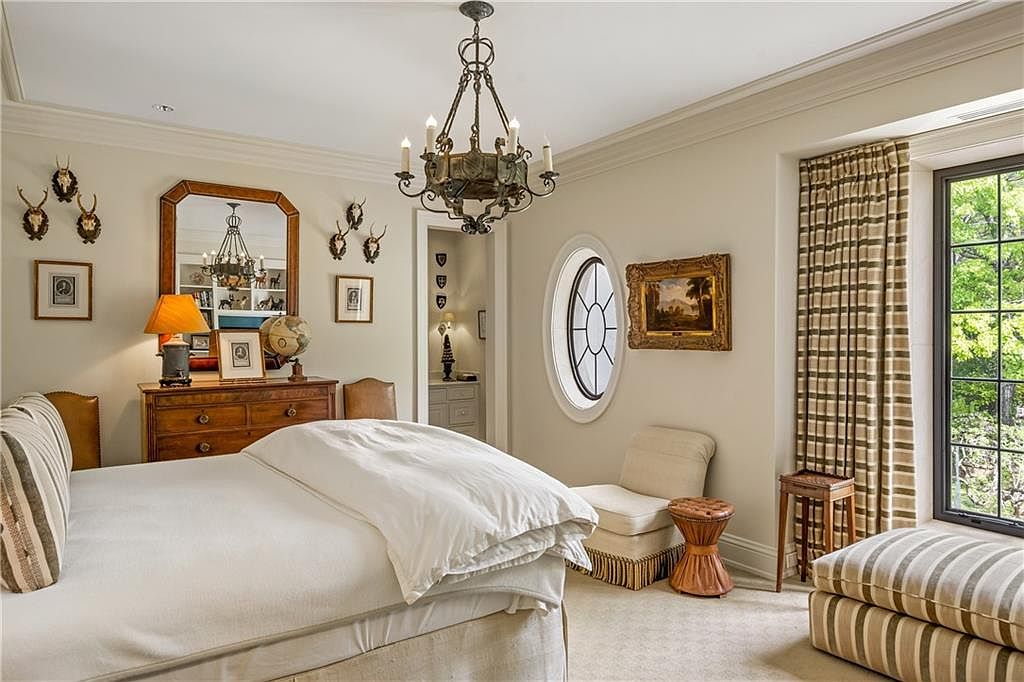 This elegant bedroom features a neutral color palette, a large bed with white linens, and a classic chandelier as a central focal point. The room includes a wooden dresser topped with a lamp and globe, a small armchair near an oval window, and a striped ottoman by the window. The space is decorated with framed artwork and wall-mounted animal decor, creating a sophisticated and traditional atmosphere.