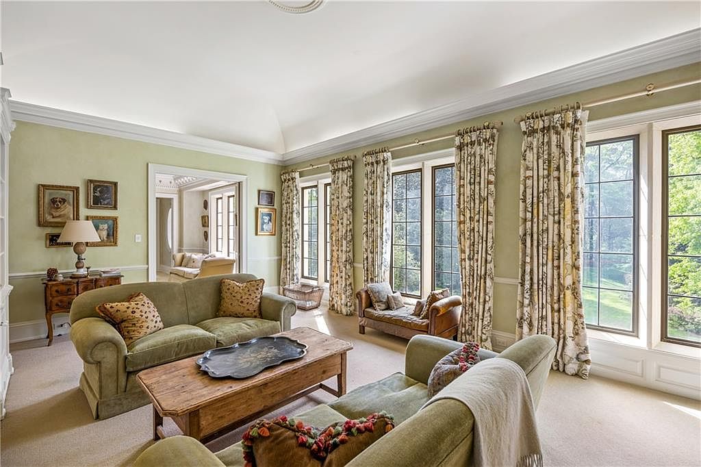 This elegant living room features soft green walls, plush carpeting, and a vaulted ceiling that enhances the sense of space. The room is furnished with two comfortable sofas, a rustic wooden coffee table, and a small leather chaise lounge positioned near the large windows. Natural light streams through the tall windows, which are dressed in patterned curtains, offering a serene view of the outdoors.