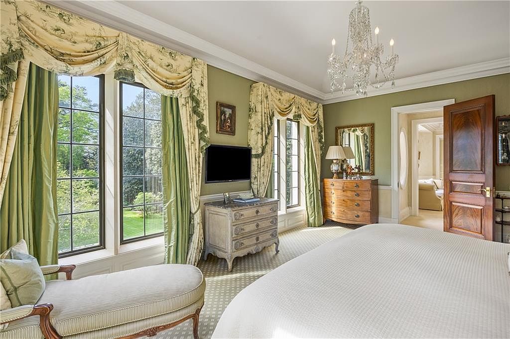 This elegant primary bedroom features a classic, traditional aesthetic with soft green walls, patterned window treatments, and a crystal chandelier. A chaise lounge sits near large windows that provide ample natural light and views of the lush exterior, while a wooden dresser serves as a media console beneath a wall-mounted television. The room flows into an adjacent space through a rich wood door, creating a sophisticated and inviting atmosphere.