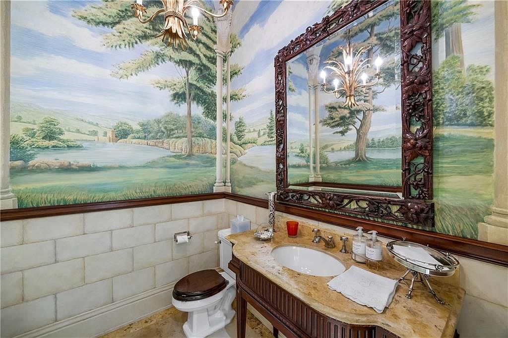 This powder room features a distinctive mural depicting a serene landscape with trees and a lake, creating an immersive, artistic atmosphere. The space is appointed with a vanity topped with a stone counter, an ornate dark wood-framed mirror, and a classic toilet with a dark wood seat. The lower walls are finished with light-colored rectangular tiles, providing a balanced contrast to the elaborate wall mural above.