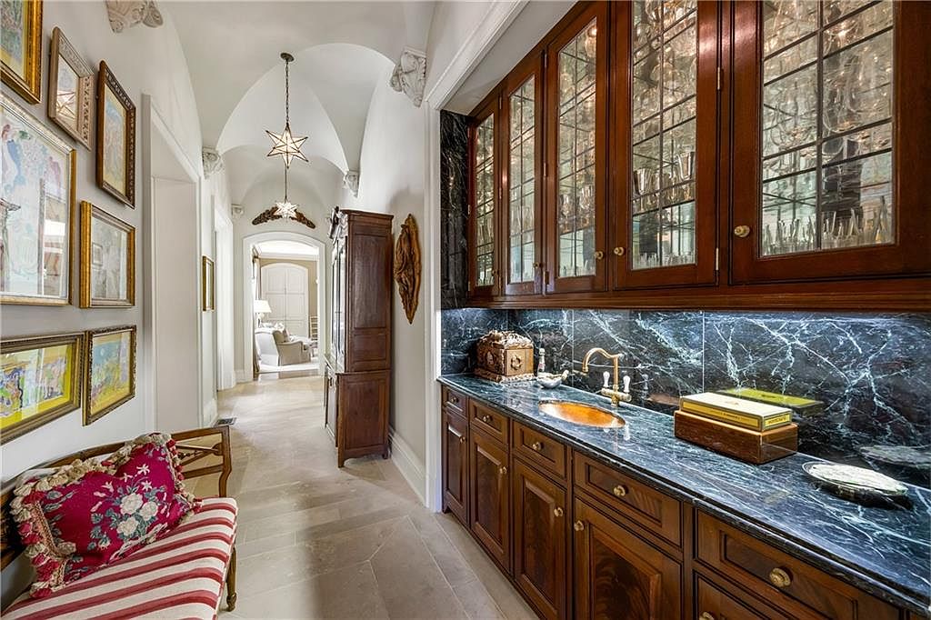 This elegant hallway features a vaulted ceiling with decorative corbels and two star-shaped pendant lights, leading toward a bright, open living space in the distance. To the right, a built-in wet bar showcases dark wood cabinetry with glass-front doors and a dark stone countertop with a matching backsplash. A striped bench with a floral pillow sits on the left, beneath a gallery wall of framed artwork, creating a sophisticated and welcoming transition space.