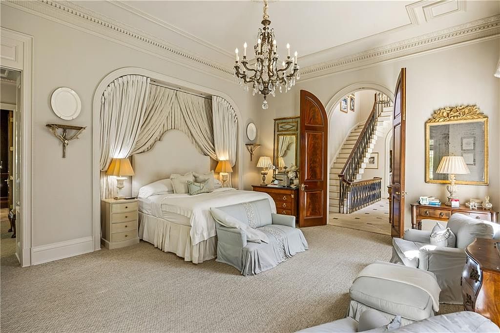 This elegant primary bedroom features a grand bed set within a recessed arched alcove draped in soft, neutral fabric curtains. The room is appointed with a crystal chandelier, plush carpeting, and a seating area, all framed by classic architectural molding and arched doorways that lead to a grand staircase. The space exudes a sophisticated, traditional aesthetic with warm lighting and high-end finishes.