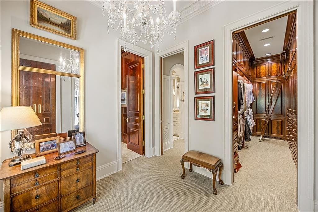 This elegant hallway features a classic design with a crystal chandelier, a wooden dresser topped with a lamp, and a small upholstered bench. The space provides access to multiple rooms, including a large walk-in closet with custom wood cabinetry and a rolling ladder. The neutral walls are adorned with framed artwork, creating a sophisticated and well-appointed transition area.