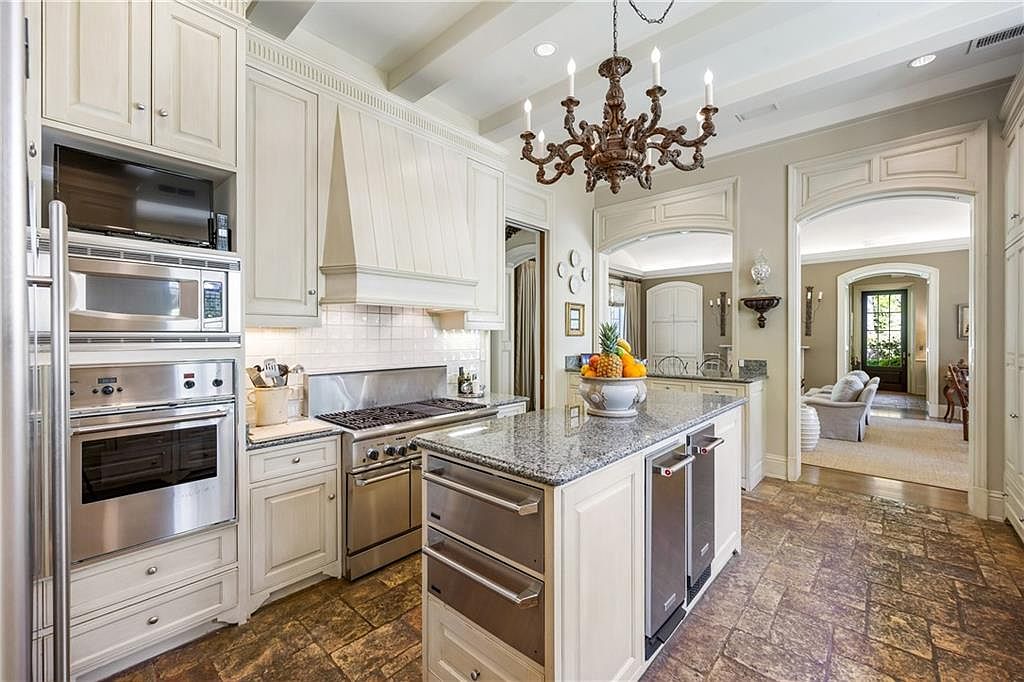 This elegant kitchen features cream-colored cabinetry, a large central island with a stone countertop, and high-end stainless steel appliances. A decorative chandelier hangs above the island, while the floor is finished in textured, earth-toned stone tiles. The space flows seamlessly into an adjacent living area through a wide, arched doorway, creating an open and inviting atmosphere.
