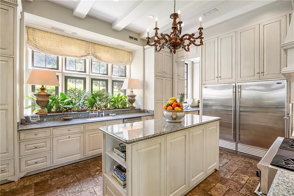 This elegant kitchen features light-toned cabinetry, a central island with a stone countertop, and a large window bank overlooking lush greenery. A decorative chandelier hangs above the island, while stainless steel appliances and textured stone flooring complete the sophisticated, traditional aesthetic. The perspective is a wide-angle shot from the kitchen entrance, showcasing the functional layout and bright, airy atmosphere.