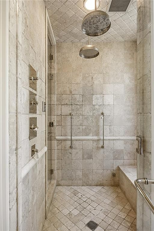 This walk-in shower features light-toned stone tile throughout, including a diamond-patterned floor and a built-in bench. The space is equipped with dual overhead showerheads and a vertical array of wall-mounted controls, all finished in a polished metallic tone. The perspective looks directly into the shower enclosure, highlighting the clean, spa-like aesthetic and the glass door entry.