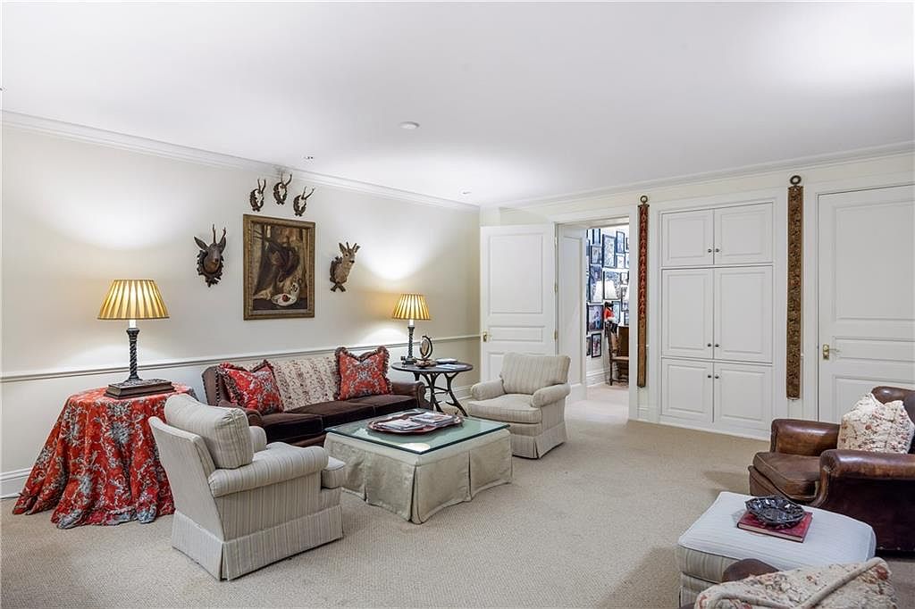 This traditional living room features a comfortable seating arrangement with a dark sofa, two light-colored armchairs, and a leather armchair, all centered around a glass-topped ottoman. The walls are adorned with framed artwork and decorative animal mounts, while a round side table and a table with a red patterned cloth add character to the space. A doorway leads to an adjacent room, and built-in white cabinetry provides ample storage, creating a cozy and well-appointed atmosphere.