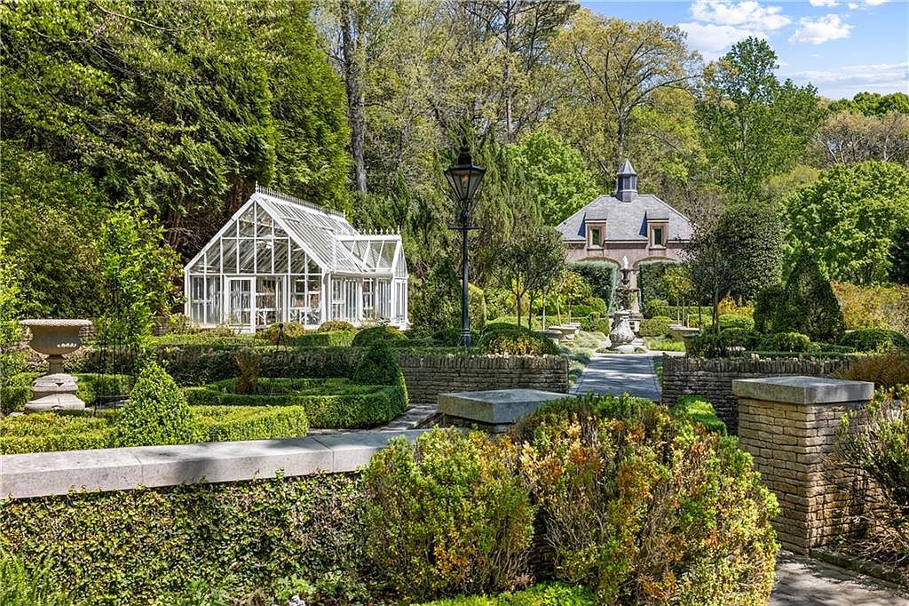 This meticulously landscaped formal garden features a white-framed glass greenhouse and a stone-built structure with a cupola in the background. The foreground is defined by manicured boxwood hedges, stone retaining walls, and a central stone fountain, all set against a backdrop of mature trees. The perspective is a wide, eye-level shot that captures the serene and elegant atmosphere of a private estate garden.
