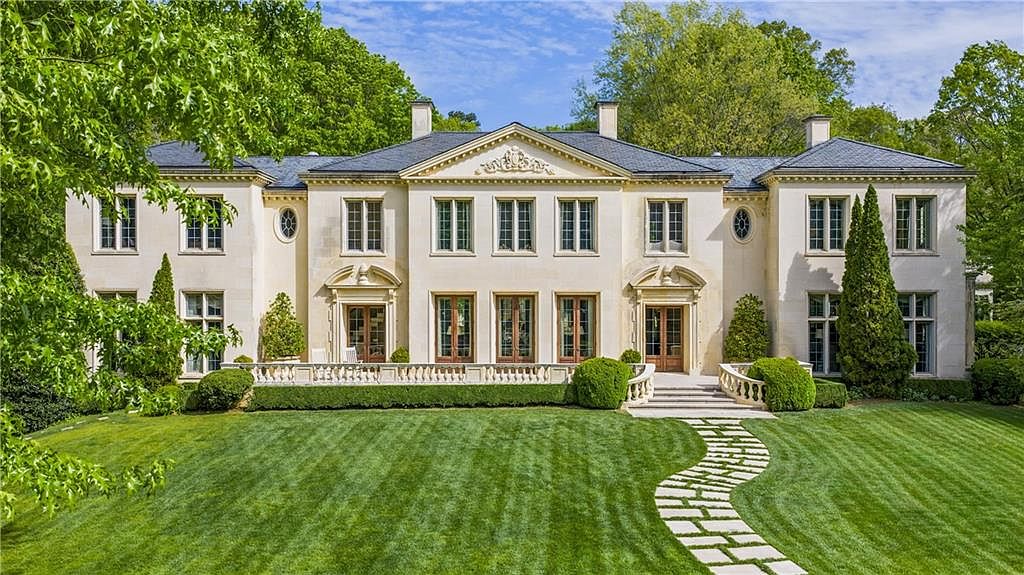 This grand, symmetrical estate features a light-colored stone facade with a classic pediment and a slate-tiled roof. A manicured lawn leads to a stone-paved walkway, framed by lush green hedges and mature trees that provide a private, park-like setting. The rear elevation showcases multiple sets of French doors opening onto a stone terrace, emphasizing an elegant and stately architectural style.