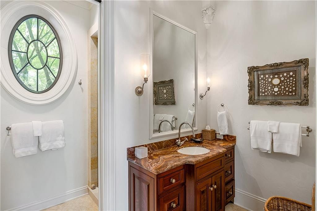 This elegant bathroom features a rich, dark wood vanity topped with a striking, multi-toned stone countertop and an integrated sink. A large, rectangular mirror is flanked by two wall-mounted light fixtures, while a unique circular window adds architectural interest to the space. The room is finished with white walls, plush white towels, and decorative framed artwork, creating a sophisticated and polished aesthetic.