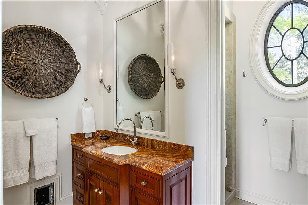 This elegant bathroom features a rich, dark wood vanity topped with a striking, heavily veined stone countertop and an undermount sink. A large, framed mirror hangs above the vanity, flanked by two wall-mounted light fixtures, while a decorative woven basket is mounted on the wall as an accent piece. To the right, a circular window with a grid pattern adds architectural interest, and white towels are neatly hung on racks, creating a clean and sophisticated aesthetic.