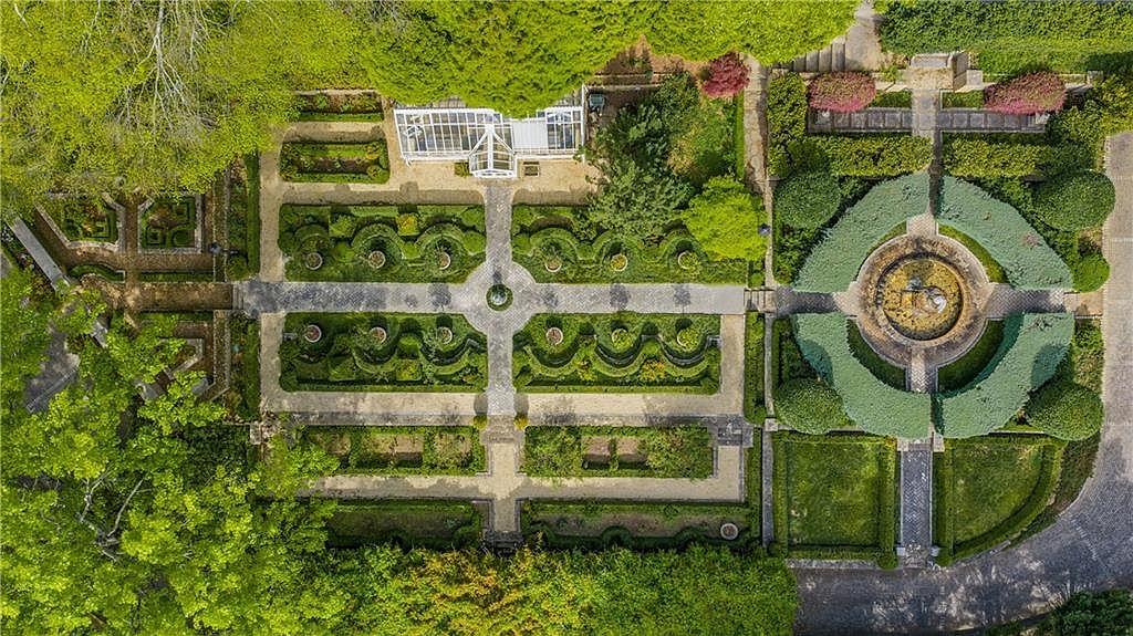 This high-angle aerial view captures a meticulously manicured formal garden featuring intricate boxwood hedges, gravel pathways, and a central circular fountain. A glass-walled conservatory sits at the top of the garden, surrounded by lush greenery and mature trees that frame the geometric landscape. The design reflects a classic, structured aesthetic, emphasizing symmetry and elegant outdoor living spaces.