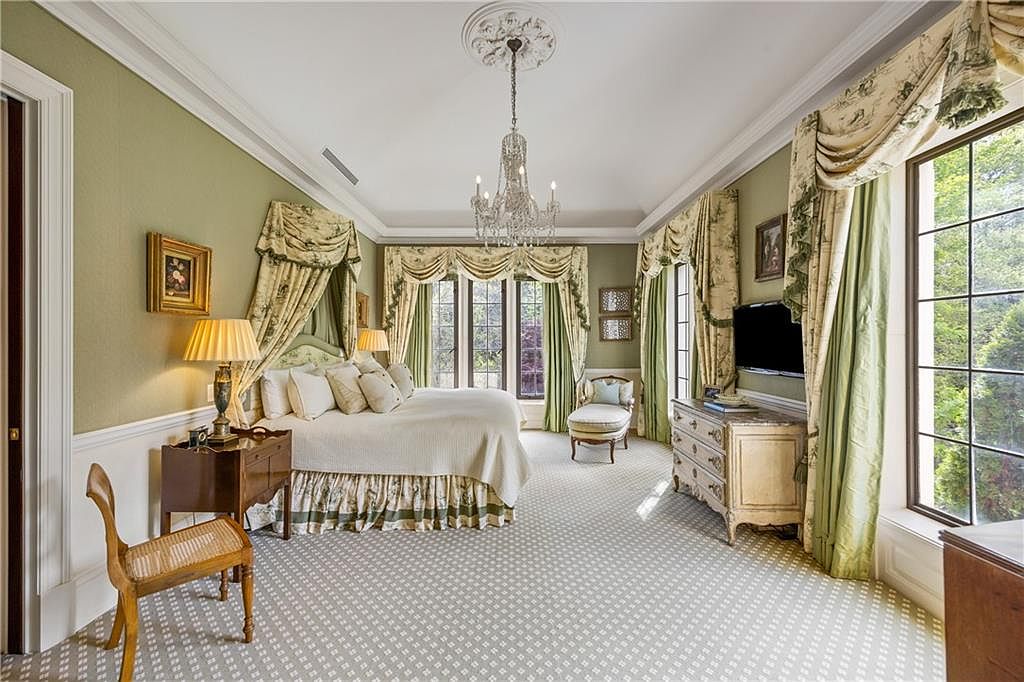 This elegant primary bedroom features a sophisticated traditional design with soft green walls, a patterned carpet, and a central chandelier hanging from a tray ceiling. A plush bed is framed by ornate fabric drapery, complemented by a wooden side table, a chaise lounge, and a classic dresser beneath a wall-mounted television. Large windows with matching valances allow ample natural light to fill the room, creating a serene and luxurious atmosphere.