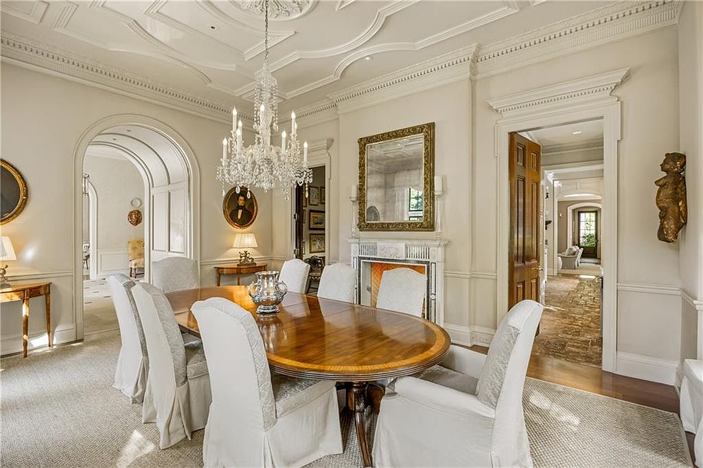 This elegant dining room features a large oval wooden table surrounded by upholstered chairs, set beneath a grand crystal chandelier. The room is characterized by sophisticated architectural details, including intricate crown molding, a fireplace with a decorative mantel, and a large gold-framed mirror. The perspective captures the formal dining space with open arched doorways leading to adjacent rooms, creating a sense of flow and luxury.
