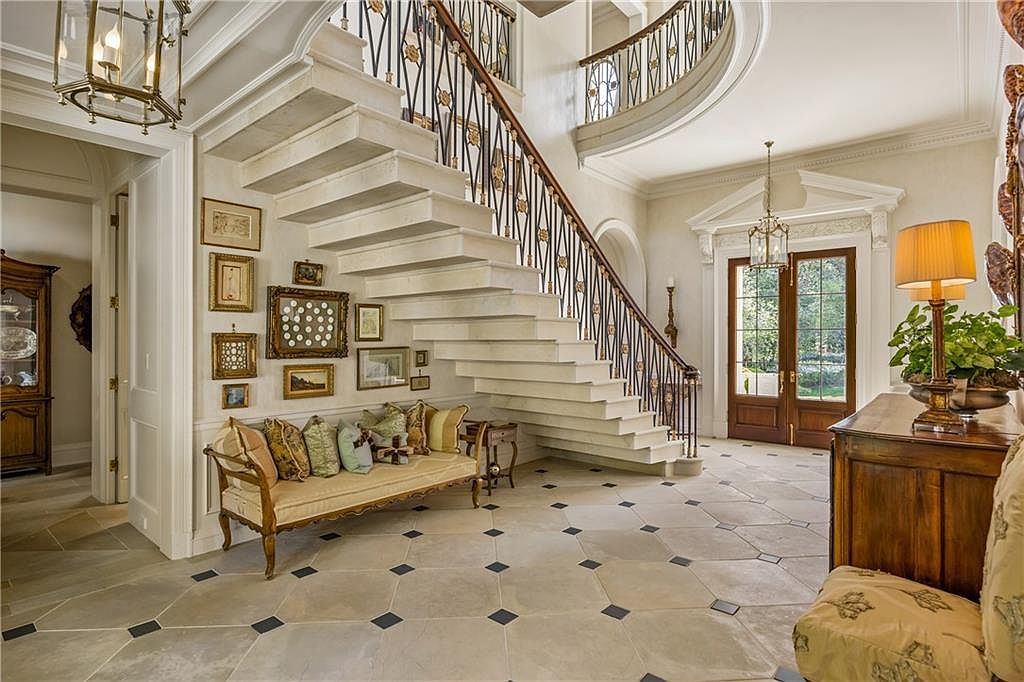 This grand foyer features a sweeping stone staircase with an intricate metal railing as its central focal point. The space is adorned with a gallery wall of framed art above a cushioned bench, set against a backdrop of light-toned walls and patterned stone flooring with dark accent inlays. A pair of glass-paned double doors at the end of the hall provides a view of the outdoors, while elegant chandeliers and a table lamp add warm, ambient lighting to the sophisticated entryway.