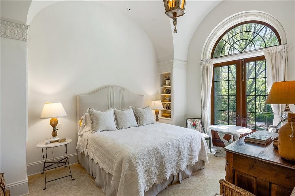 This elegant bedroom features a high, arched ceiling and a large, arched glass door that provides abundant natural light and a view of the outdoors. The room is furnished with a plush bed, a small side table with a lamp, and a wooden dresser, all set against a neutral color palette. A built-in shelving unit is recessed into the wall, adding a sophisticated architectural detail to the space.