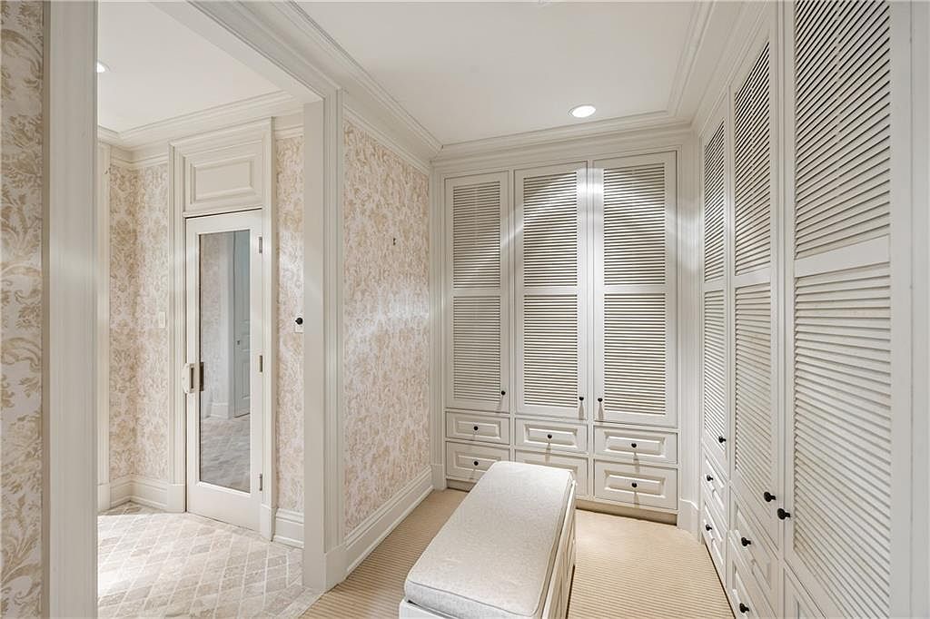 This elegant walk-in closet features extensive custom cabinetry with louvered doors and integrated drawers, providing ample organized storage. A central upholstered bench sits on the carpeted floor, while the walls are adorned with a subtle, light-toned patterned wallpaper. The space is brightened by recessed lighting and connects to an adjacent room through a glass-paned door, creating a sophisticated and airy atmosphere.