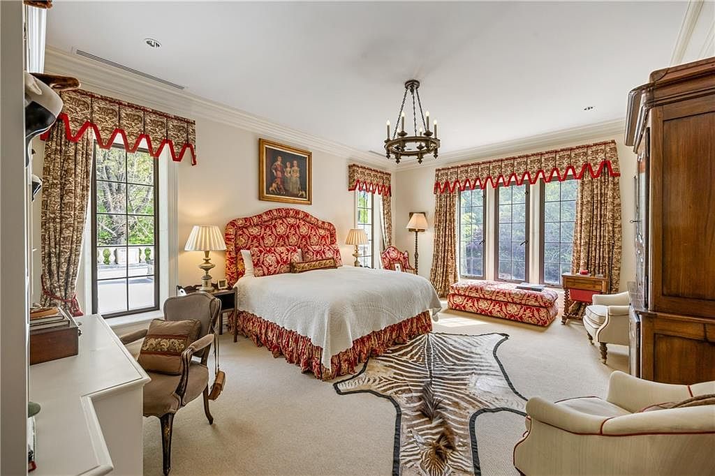 This elegant primary bedroom features a grand bed with a patterned red and gold upholstered headboard, complemented by matching window treatments and a bench at the foot of the bed. The room is anchored by a zebra-print area rug on neutral carpeting and illuminated by a classic chandelier and bedside lamps. Large windows provide ample natural light, creating a sophisticated and traditional atmosphere.