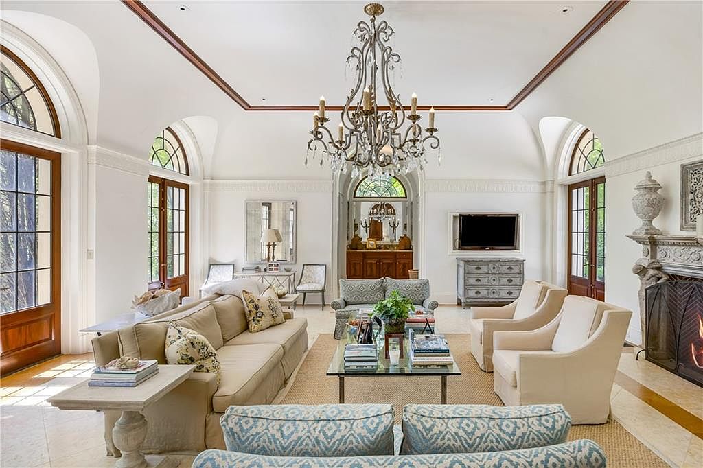 This elegant living room features a high, coved ceiling with a prominent iron chandelier and arched glass doors that provide abundant natural light. The space is anchored by a neutral-toned sofa and matching armchairs arranged around a glass-topped coffee table, with a stone fireplace surround visible on the right. The room exudes a sophisticated, classic aesthetic with its light-colored tile flooring and symmetrical layout.