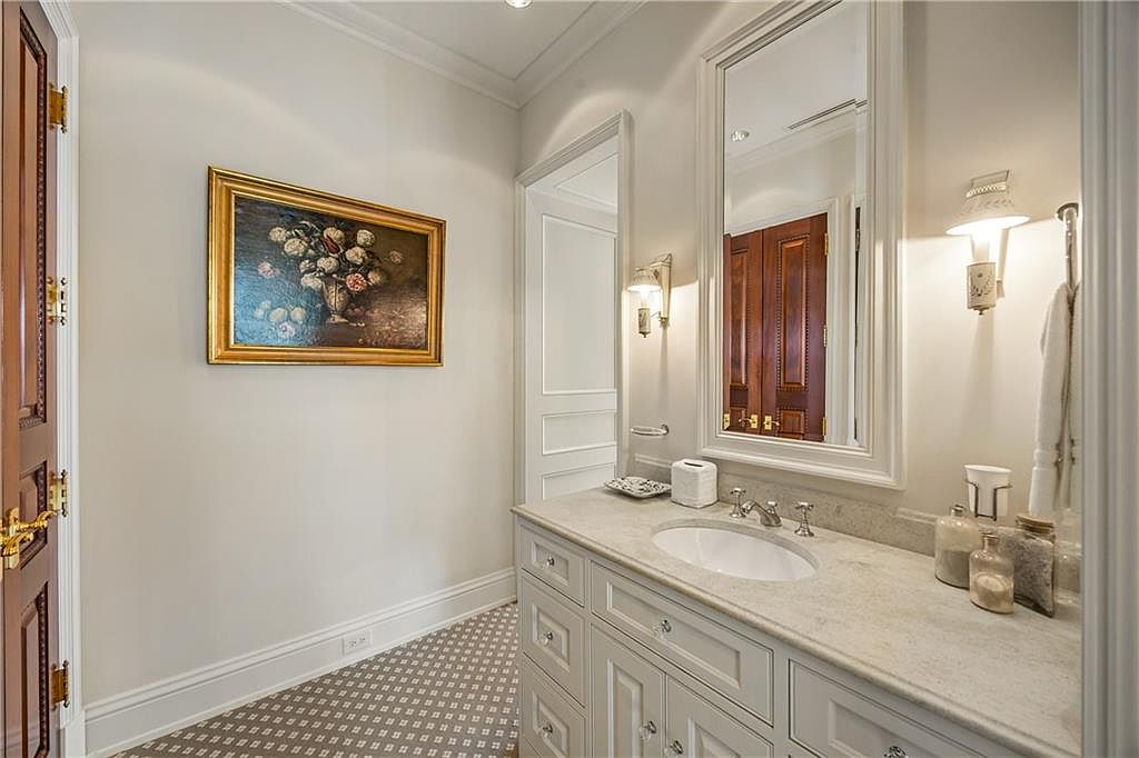 This elegant powder room features a light-toned vanity with a stone countertop and an undermount sink, complemented by a large, framed mirror. The space is adorned with a classic floral still-life painting on the wall and patterned tile flooring, creating a sophisticated and refined atmosphere. The perspective captures the vanity area and a glimpse into an adjacent room through a doorway, highlighting the home's cohesive design.