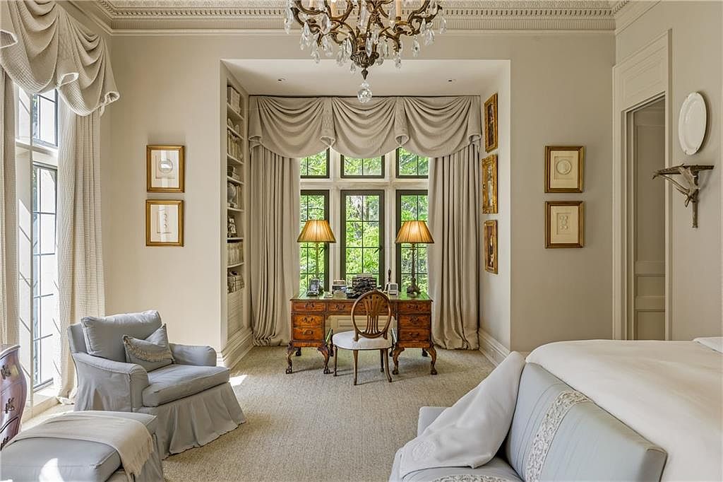 This elegant primary bedroom features a sophisticated neutral color palette, highlighted by a classic chandelier and custom window treatments framing a dedicated desk area. The room includes a comfortable seating area with a plush armchair and a portion of a bed, creating a serene and refined atmosphere. The space is well-appointed with built-in shelving, framed artwork, and a traditional desk, offering a blend of luxury and functionality.