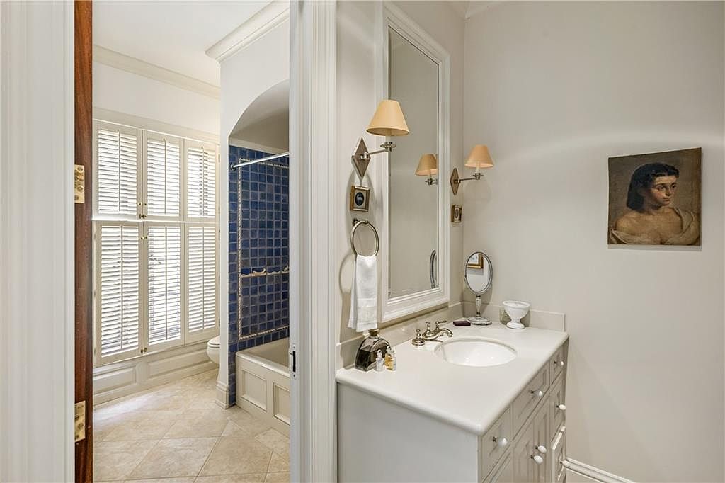 This interior view captures a well-appointed bathroom featuring a white vanity with a sink and a framed mirror flanked by wall-mounted light fixtures. To the left, an open doorway reveals a secondary space with a bathtub surrounded by blue square tiles and large, shuttered windows. The room is decorated with a framed portrait on the wall, creating a classic and refined aesthetic.