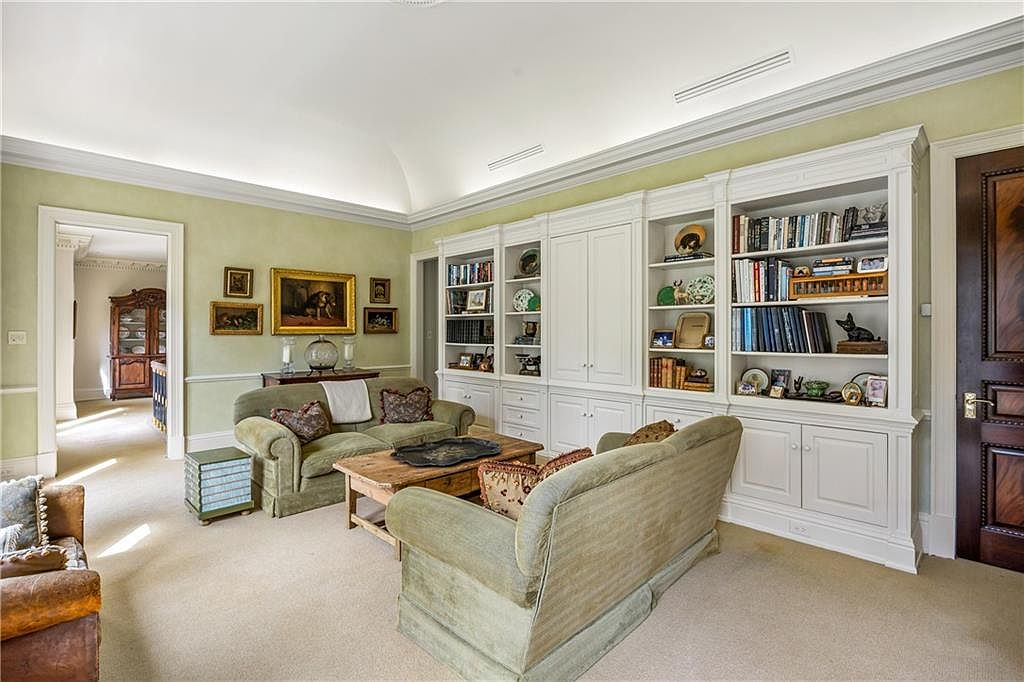 This elegant living room features a soft, neutral color palette with sage green walls and plush, light-toned carpeting. A large, custom-built white shelving unit spans the wall, providing ample display space for books and decor, while two comfortable green sofas are arranged around a rustic wooden coffee table. The room is bathed in soft, indirect lighting from the vaulted ceiling, creating a warm and inviting atmosphere that feels both traditional and sophisticated.