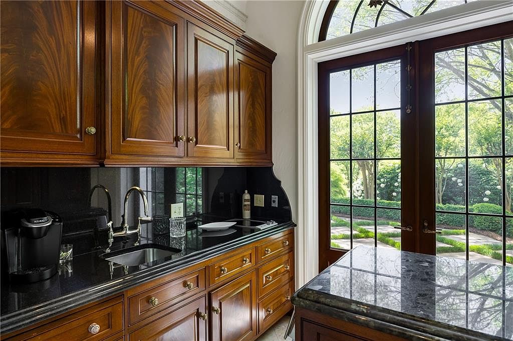 This elegant butler's pantry or wet bar area features rich, dark-stained wood cabinetry with intricate grain patterns and classic hardware. A polished black stone countertop houses an undermount sink and a modern faucet, complemented by a sleek black backsplash. To the right, tall, multi-paned glass doors provide a view of a lush, green garden, while a portion of a stone-topped island is visible in the foreground.