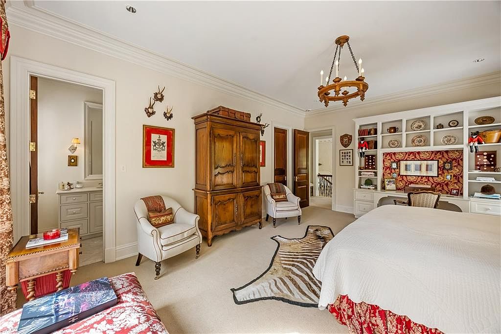 This spacious primary bedroom features a traditional design with a large wooden armoire, two elegant armchairs, and a built-in shelving unit surrounding a desk area. A zebra-print rug sits on the carpeted floor near the bed, which is dressed in white linens and a patterned red bed skirt. The room is accented with wall-mounted decor and a classic chandelier, creating a warm and sophisticated atmosphere.