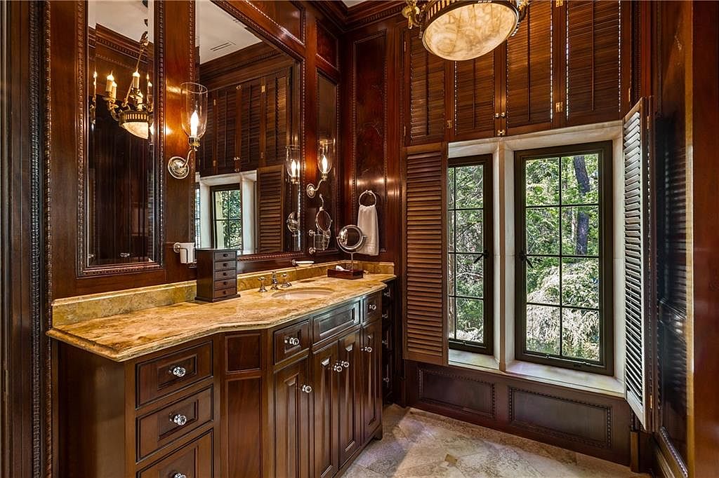This elegant bathroom features rich, dark wood cabinetry paired with a light-toned stone countertop and a large, ornate mirror. The space is framed by dark wood paneling and shutters that cover the windows, which look out onto a lush, green forest view. A decorative chandelier hangs from the ceiling, adding a touch of luxury to the warm, traditional aesthetic.
