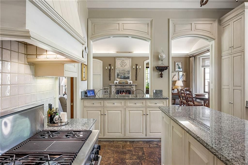 This elegant kitchen features light-toned cabinetry, granite countertops, and a professional-grade gas range. The space is defined by two large arched openings that provide a clear view into an adjacent living area with a fireplace, creating an open and airy atmosphere. The perspective from the kitchen island highlights the seamless transition between cooking and living spaces, emphasizing the home's sophisticated, traditional design.