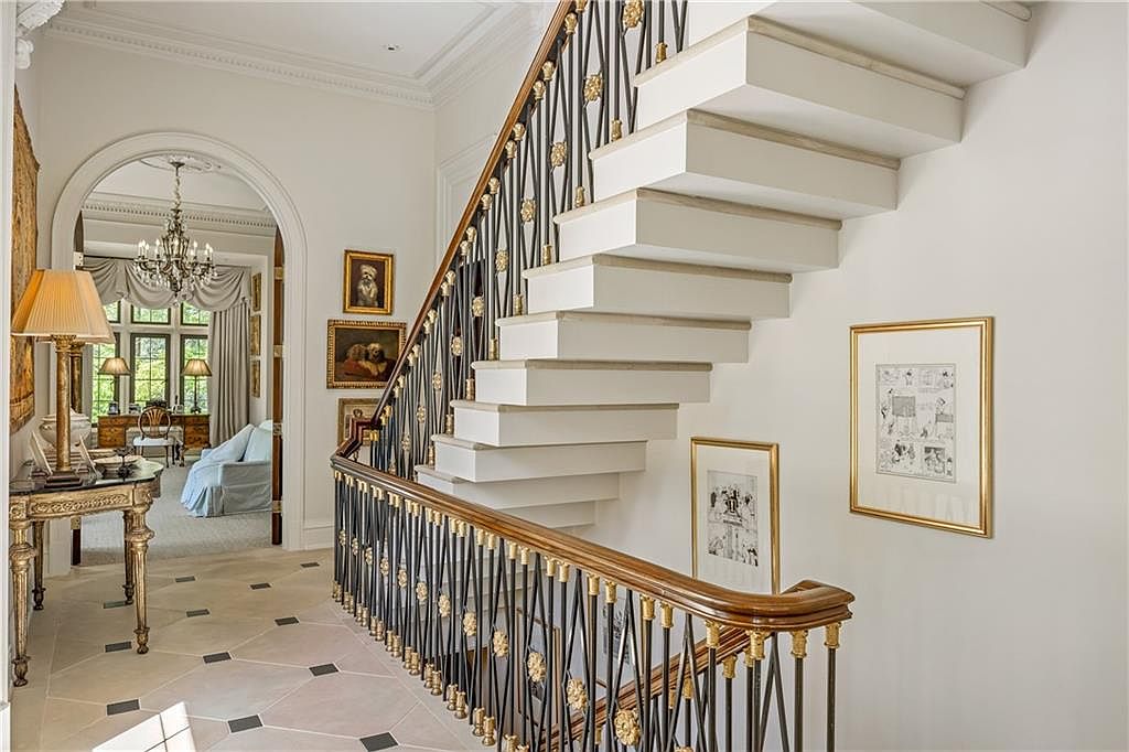 This elegant foyer features a striking floating staircase with white treads and a decorative black metal railing accented with gold-toned floral motifs. To the left, an arched doorway leads into a bright living area, while the floor is finished in light-colored stone tiles with dark inlay accents. The space is adorned with framed artwork and a classic console table, creating a sophisticated and welcoming atmosphere.