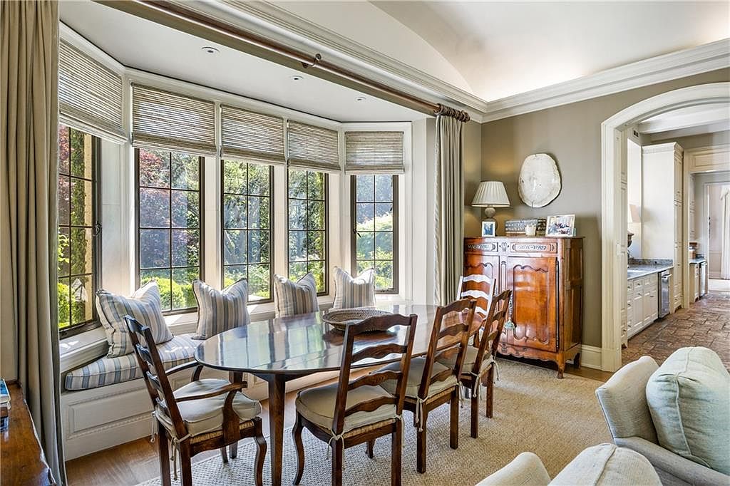 This elegant dining room features a large oval wooden table surrounded by classic chairs, set against a bright bay window with built-in bench seating. A traditional wooden sideboard stands against the wall, topped with a decorative plate and a table lamp, while a doorway leads into a bright, tiled kitchen area. The room is bathed in natural light, creating a warm and inviting atmosphere suitable for formal or casual gatherings.