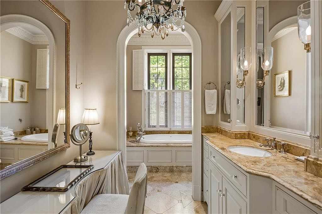 This elegant primary bathroom features a dual-vanity area with stone countertops and a dedicated makeup vanity with a skirted base. An arched doorway leads to a separate bathing area containing a soaking tub positioned beneath a window with plantation shutters. The space is finished with neutral tones, a crystal chandelier, and polished stone flooring, creating a sophisticated and tranquil atmosphere.