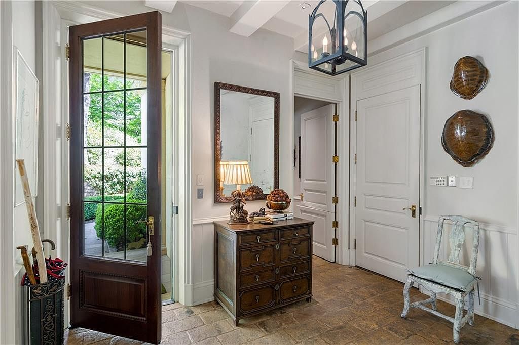 This elegant entryway features a dark wood-framed glass door that opens to a lush outdoor view, creating a bright and welcoming transition. The space is furnished with a classic wooden chest topped with a mirror and lamp, complemented by a distressed-finish chair and decorative wall-mounted turtle shells. The stone-tiled floor and neutral wall tones contribute to a sophisticated, traditional aesthetic.