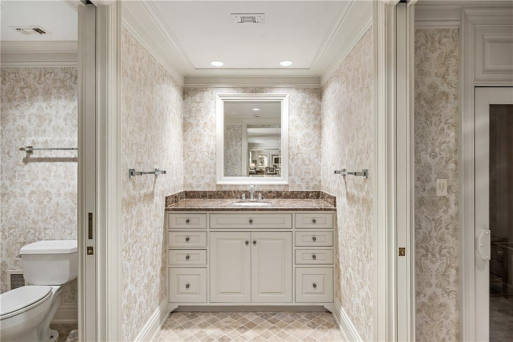 This elegant bathroom features a central vanity with a stone countertop, a framed mirror, and classic cabinetry. The walls are adorned with a sophisticated damask-patterned wallpaper, complemented by neutral-toned floor tiles. The space is framed by white trim and doorways, creating a clean and traditional aesthetic.