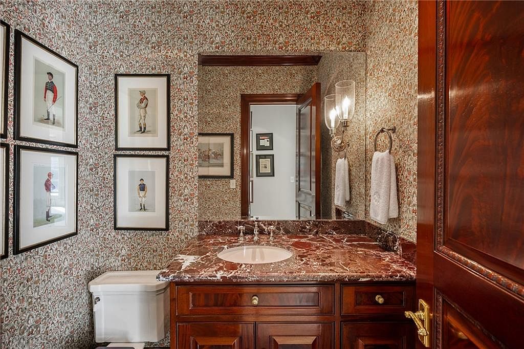 This traditional powder room features ornate, patterned wallpaper and a rich, dark wood vanity topped with a polished stone surface. A large mirror reflects the space, including a wall-mounted light fixture and framed equestrian-themed artwork that adds a classic, sophisticated touch. The room is framed by a dark wood door, creating a cohesive and elegant aesthetic.