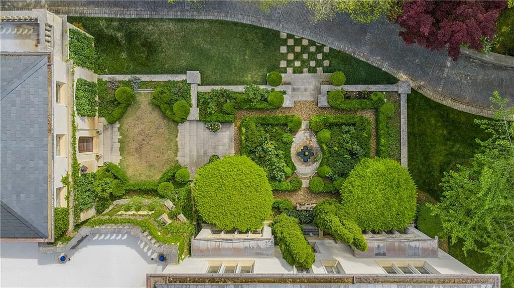This high-angle aerial view captures a meticulously landscaped formal garden nestled between two wings of a grand estate. The garden features geometric boxwood hedges, a central stone fountain, and a mix of manicured lawns and gravel pathways. The perspective highlights the architectural symmetry of the building's rooflines and the lush, mature greenery surrounding the property.