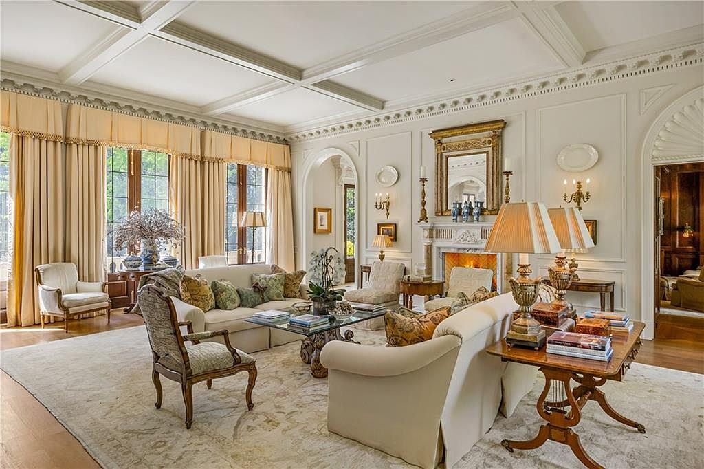 This elegant, formal living room features a coffered ceiling, intricate crown molding, and large windows draped in floor-length curtains. A central fireplace with a decorative mantel serves as the focal point, flanked by wall sconces and framed by a symmetrical furniture arrangement including plush sofas and armchairs. The space is anchored by a large, neutral-toned area rug over hardwood floors, creating a sophisticated and inviting atmosphere.