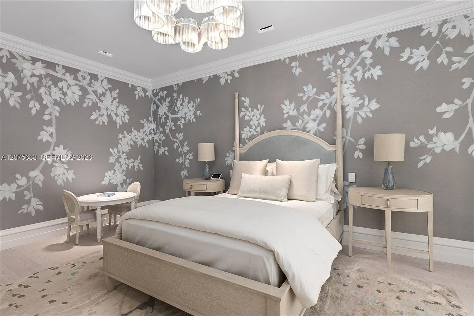 This elegant bedroom features a sophisticated gray wall treatment adorned with a delicate white floral pattern, creating a serene and artistic atmosphere. The room is anchored by a light-toned wooden bed frame and matching nightstands, complemented by a unique, multi-tiered glass chandelier that serves as a focal point. A small, circular table with two chairs in the corner adds a charming touch, while the soft, neutral color palette and light wood flooring provide a cohesive and inviting aesthetic.