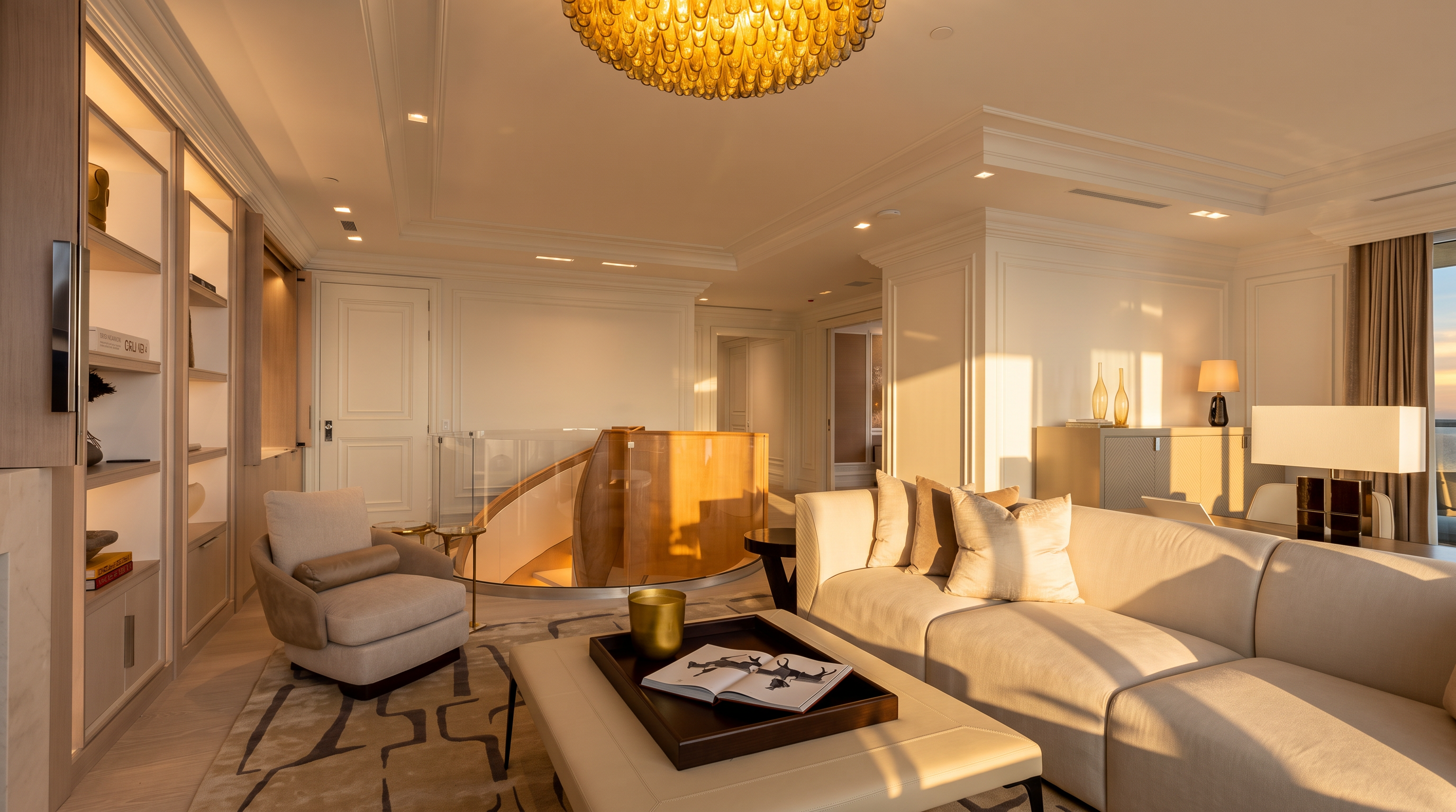 This sophisticated living room features a neutral color palette, a plush white sofa, and a comfortable armchair arranged around a central ottoman with a decorative tray. A prominent curved staircase with a glass railing serves as a striking architectural focal point, while a large, textured gold chandelier hangs from the ceiling. The space is accented by built-in shelving and elegant wall molding, creating a refined and luxurious atmosphere.