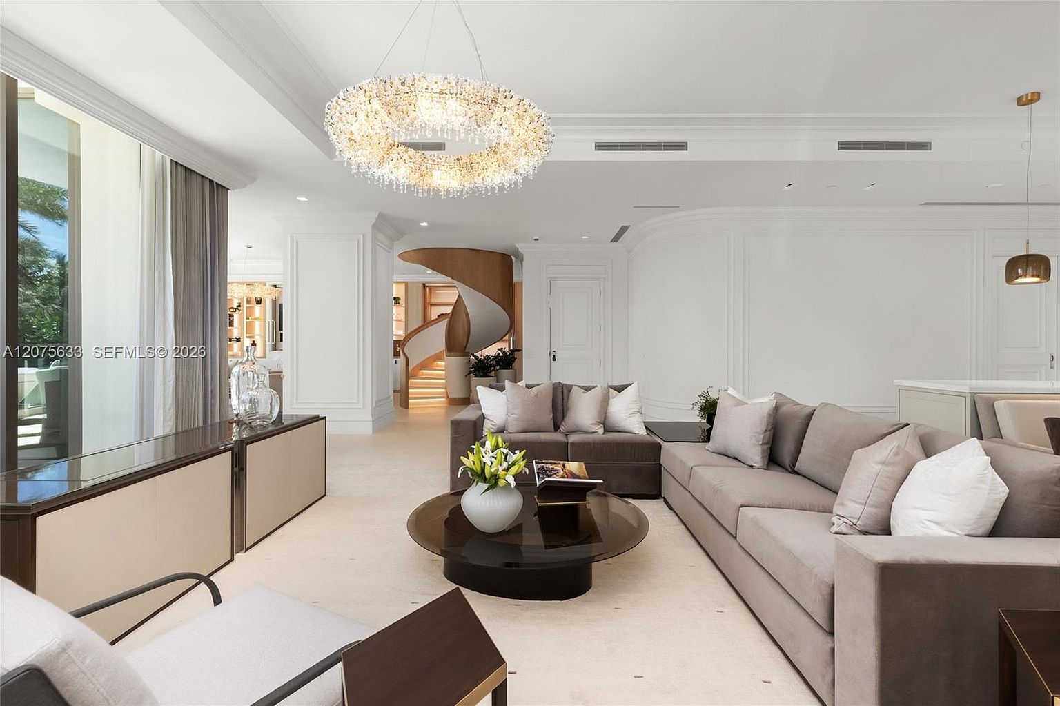 This elegant, open-concept living room features a neutral color palette and a sophisticated, modern aesthetic. A large, circular crystal chandelier serves as a focal point above the seating area, which includes a plush grey sofa and a dark, circular coffee table. In the background, a striking curved staircase with light wood accents adds architectural interest, while large windows to the left allow for ample natural light.