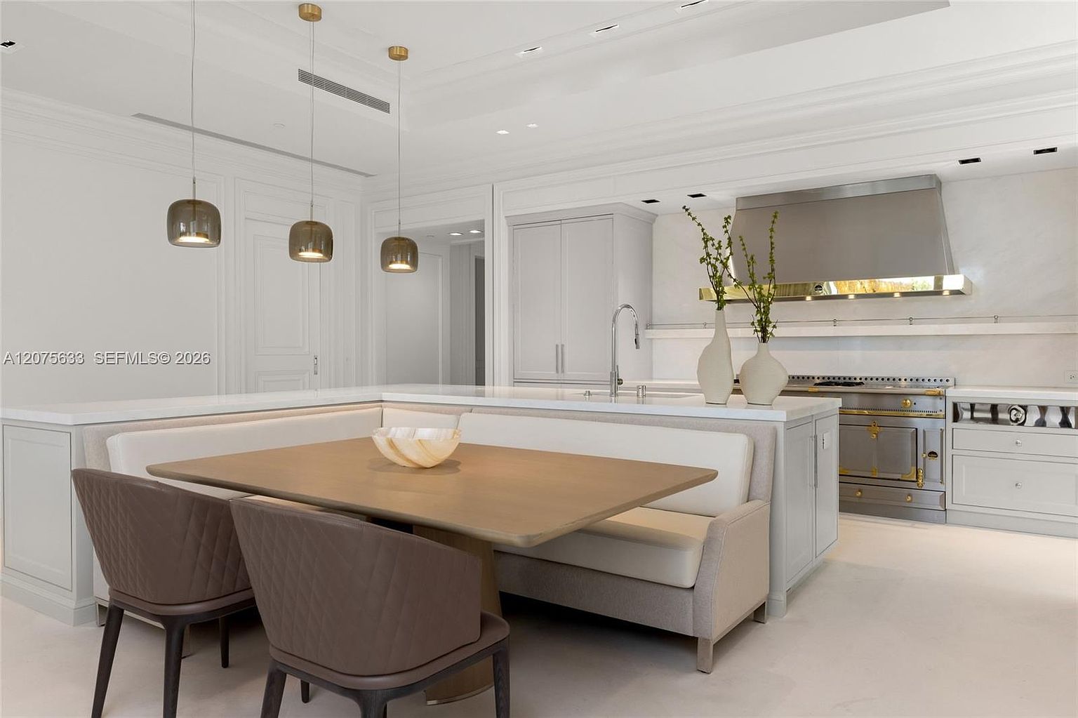 This sophisticated kitchen features a seamless blend of light-toned cabinetry and a large central island that incorporates a built-in dining banquette. The space is illuminated by three modern pendant lights hanging above the dining area, while a professional-grade stainless steel range and hood serve as the focal point of the cooking zone. The overall aesthetic is bright, airy, and highly refined, showcasing a clean, contemporary design with high-end finishes.