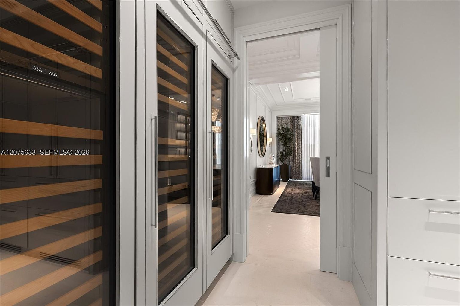 This image captures a sophisticated wine storage area featuring tall, glass-fronted wine refrigerators with light wood shelving. The perspective looks through a doorway into an adjacent dining area, which is decorated with a large circular mirror, a dark console table, and a potted plant. The space is characterized by a clean, neutral color palette and elegant, modern architectural details.