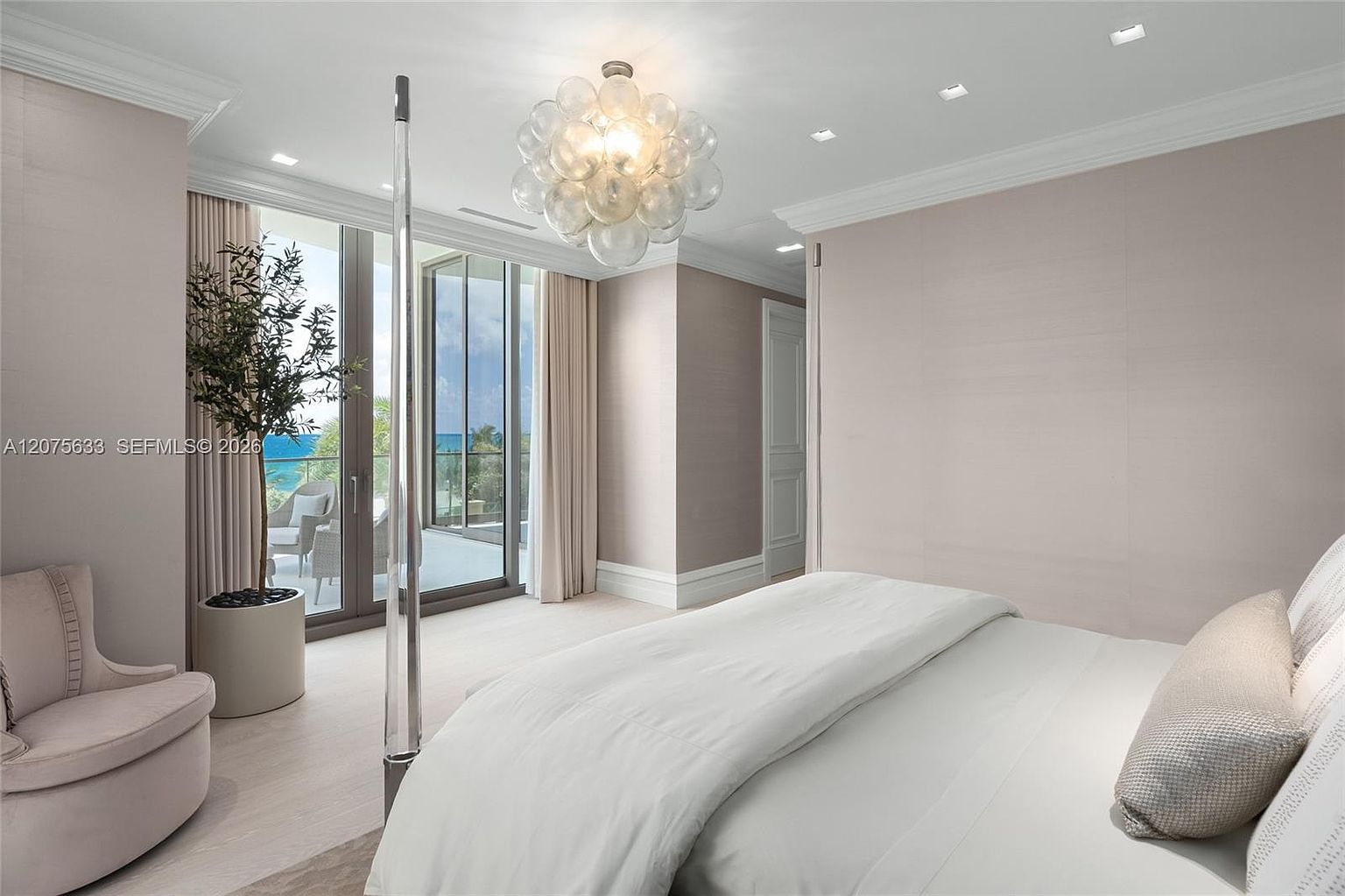 This elegant primary bedroom features a neutral color palette with soft blush walls and plush white bedding. A striking, multi-globe glass chandelier serves as a focal point above the bed, while floor-to-ceiling glass doors provide seamless access to a private balcony with ocean views. A potted tree adds a touch of nature to the serene, sophisticated space.