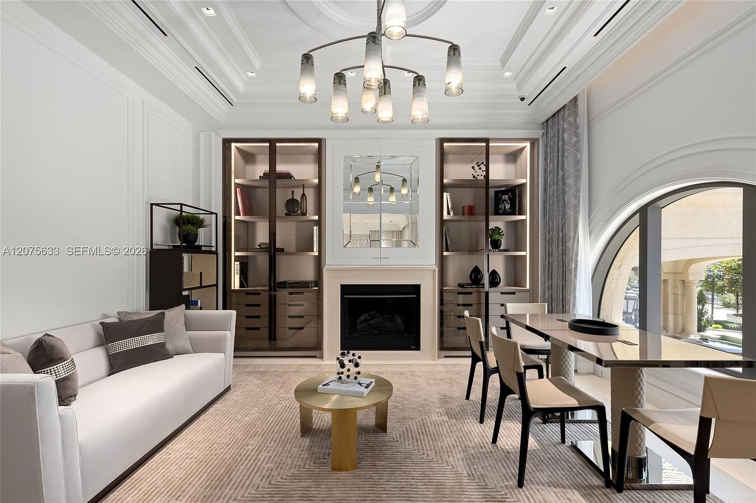 This sophisticated living room features a symmetrical layout centered around a fireplace with a decorative mirror above it, flanked by two tall, glass-fronted display cabinets. A plush white sofa sits opposite a dining area with a sleek table and modern chairs, all set upon a textured area rug. The room is illuminated by a contemporary multi-arm chandelier and benefits from natural light streaming through a large, arched window on the right.