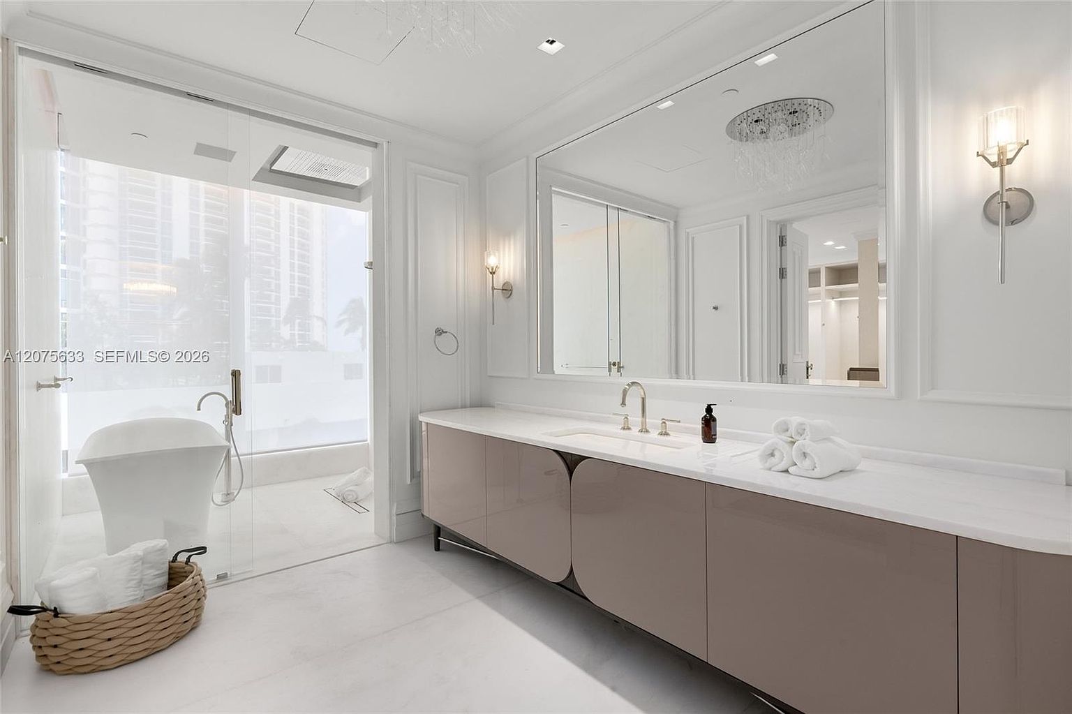 This luxurious primary bathroom features a sleek, modern vanity with a long stone countertop and a large, wall-spanning mirror. To the left, a glass-enclosed wet room houses a freestanding soaking tub, while a woven basket with towels adds a touch of texture to the minimalist white space. The room is bright and airy, with clean lines and contemporary lighting fixtures that create a sophisticated, spa-like atmosphere.