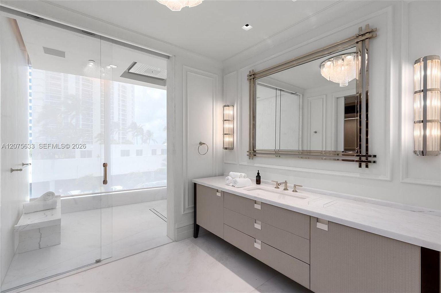 This luxurious primary bathroom features a sleek, light-toned vanity with a stone countertop and a large, ornate rectangular mirror flanked by vertical wall sconces. To the left, a glass-enclosed shower area includes a built-in stone bench and a view through a large window. The space is finished with light-colored flooring and elegant wall molding, creating a bright and sophisticated atmosphere.