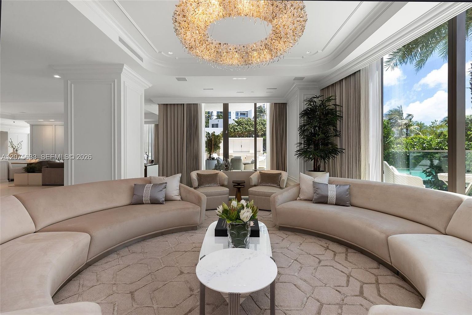 This elegant living room features a symmetrical layout with two large, curved beige sofas facing each other around a central coffee table. A striking, circular crystal chandelier serves as the focal point above the seating area, while expansive windows provide abundant natural light and views of the lush outdoor greenery. The space is finished with a neutral, textured area rug and sophisticated decor, creating a luxurious and inviting atmosphere.