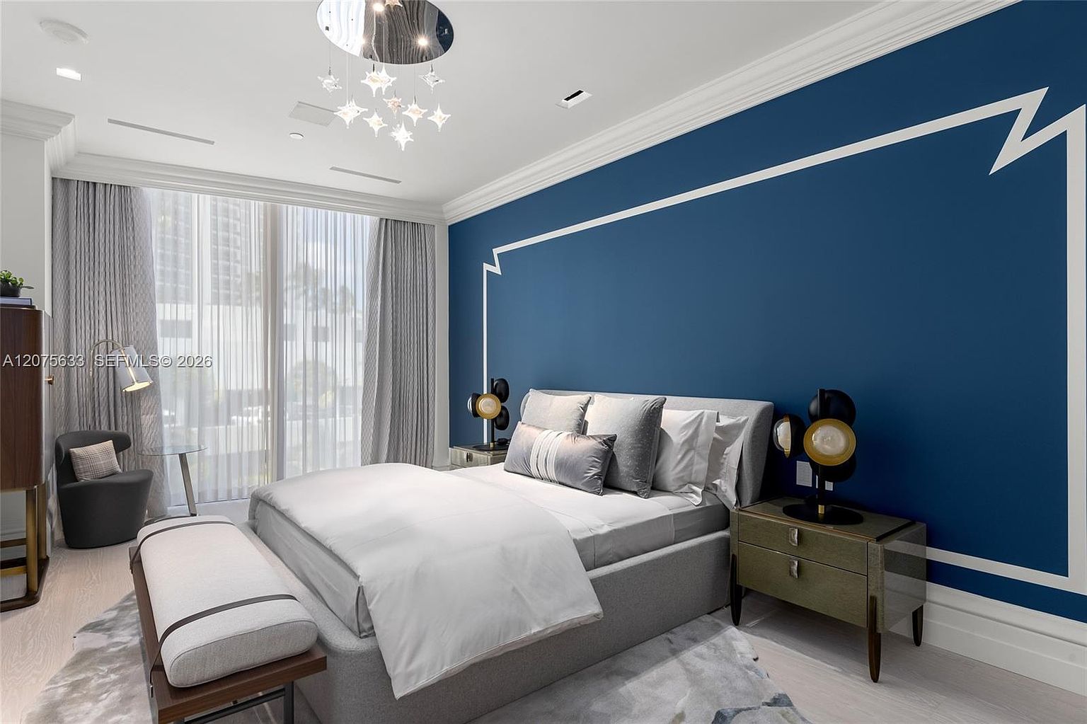 This modern primary bedroom features a bold navy blue accent wall with white decorative molding, creating a sophisticated focal point behind the upholstered bed. The room is bathed in natural light from large windows draped in floor-to-ceiling sheer curtains, while a unique star-shaped chandelier adds a touch of elegance to the space. A plush area rug, a sleek bench at the foot of the bed, and contemporary bedside lamps complete the refined, high-end aesthetic.